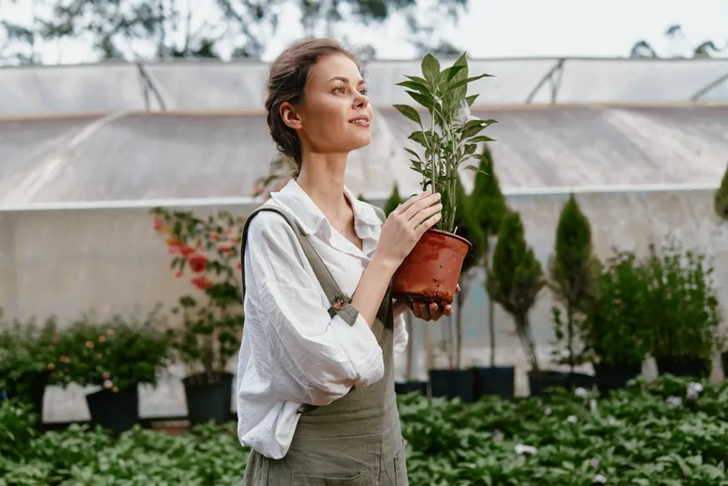 Young woman with potted plant in a greenhouse, wearing casual outfit, embracing nature, joyful expression, green plants in the background, eco friendly lifestyle