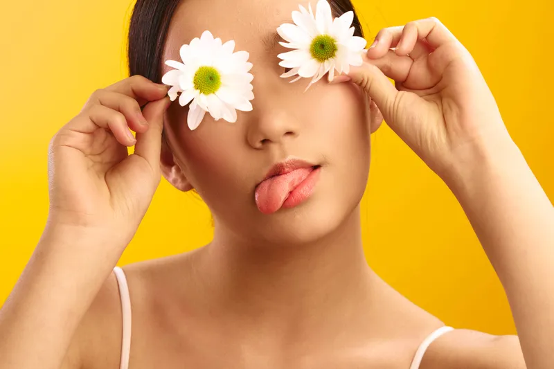 Asian woman playful poses with white flowers held over her eyes, tongue out, against a bright yellow backdrop, conveying whimsy, freshness, and creative energy.