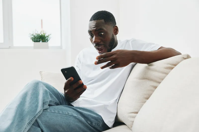 Engaged young Black man sitting on a sofa, using a smartphone with a surprised expression, showcasing modern lifestyle and technology interaction
