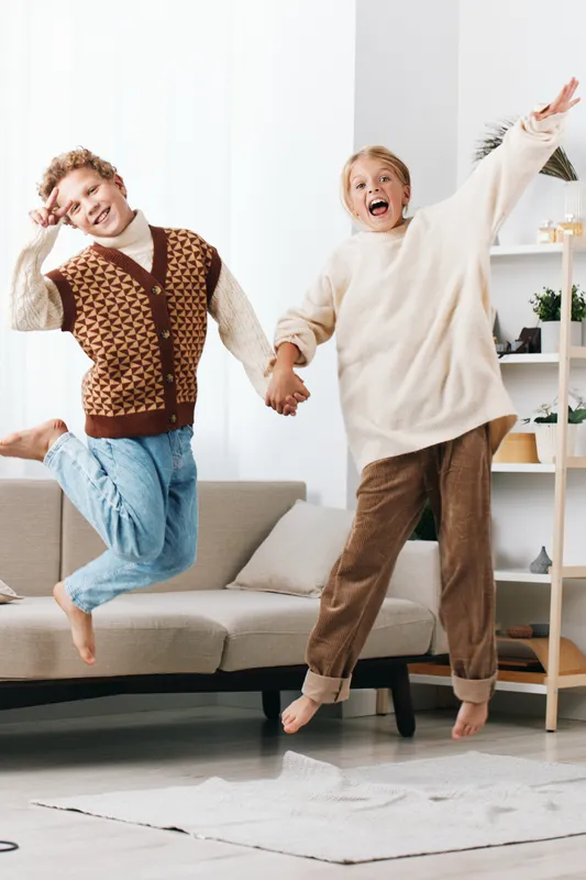 Two friends leap joyfully indoors, holding hands, wearing cozy sweaters in a bright living room. Laughter and movement capture a spontaneous moment of friendship and energy.