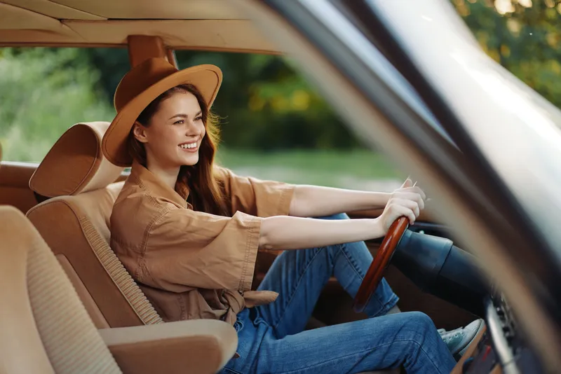 Young woman with long hair wearing a brown hat, smiling joyfully while driving an old car in a sunny outdoor setting, capturing a sense of adventure and freedom.