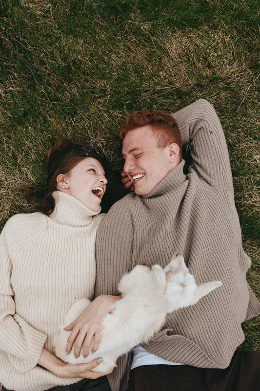 Couple lying on grass, wearing cozy sweaters, smiling at each other, with a fluffy white dog between them, enjoying a relaxed outdoor moment and warm connection, intimate lifestyle scene on a sunny
