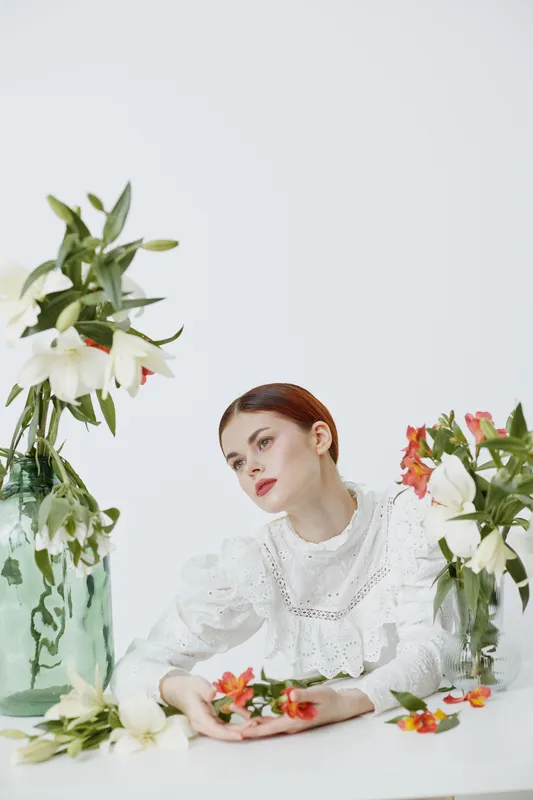 Elegant woman in white dress surrounded by fresh flowers, showcasing beauty and tranquility in a bright, minimalistic setting