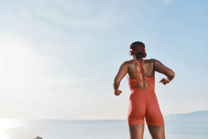 Подтянутая женщина в ярком оранжевом купальнике бежит вдоль береговой линии, наслаждаясь солнечной погодой и океанским бризом