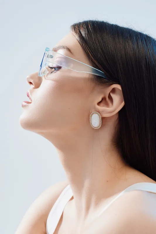 Profile of a woman with clear skin wearing transparent glasses and a white earring. Soft lighting highlights her beauty and fashion style in a minimalistic studio setting.