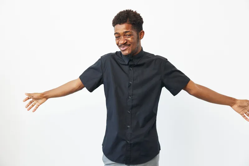 Smiling black man with open arms, expressing joy and warmth, dressed in a casual black shirt, standing against a white background.