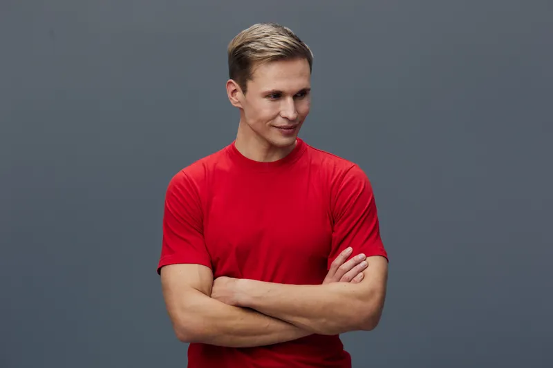 Smiling young man with blond hair, wearing a red t-shirt, arms crossed, looking to the side isolated on plain gray background. Casual fashion portrait. People lifestyle concept in studio setting.