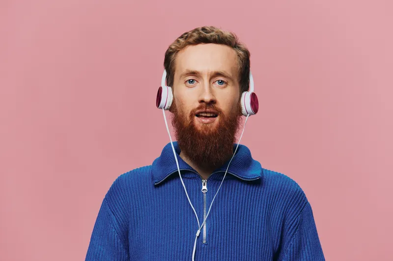 Портрет рыжеволосого мужчины в наушниках, который улыбается и танцует, слушая музыку на розовом фоне