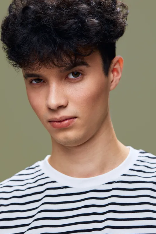 young man with curly hair wearing striped shirt posing confidently with subtle smile and neutral background, natural light enhancing facial features and warm skin tone