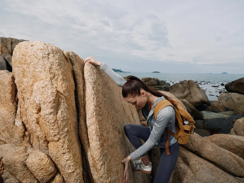Young woman climbing rocky terrain by the sea, showcasing determination and adventure in outdoor fashion with a backpack, perfect for travel and fitness enthusiasts