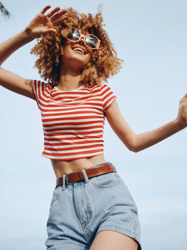 Joyful young woman dances outdoors in bright sunlight, wearing a striped crop top, denim shorts, and round sunglasses, capturing carefree summer energy and movement.