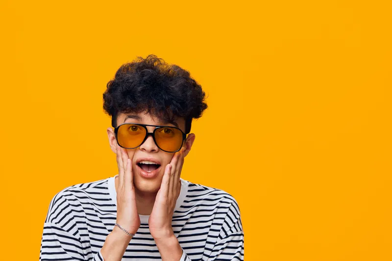 surprised young man wearing striped shirt and yellow sunglasses with curly hair on bright orange background, expressing shock and excitement.