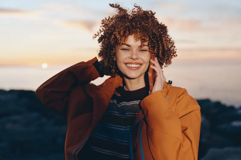 Woman with curly hair smiling outdoors at sunset, orange jacket, phone near her ear, warm lifestyle portrait, soft light, clear focus, joyful mood