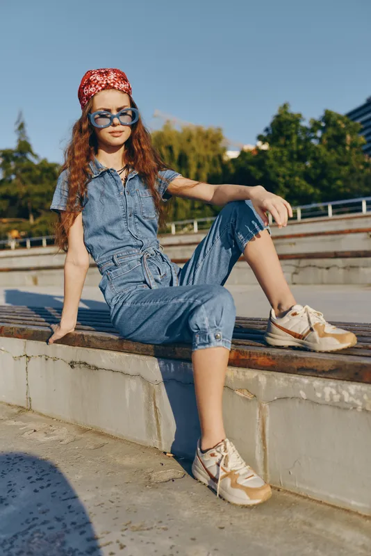 teenage girl wearing denim jumpsuit and sneakers relaxing outdoors in summer, with sunglasses and red bandana on sunny day