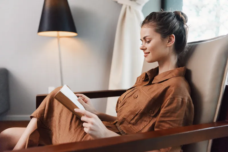 Умиротворённая молодая женщина наслаждается чтением книги в своей современной гостиной в тёплых тонах, залитой естественным светом и окружённой стильным декором