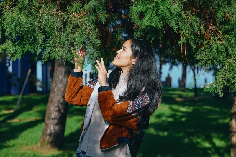 Woman near hanging greenery among trees in a sunny park, exploring nature and foliage with curiosity and joy, light breeze and carefree atmosphere