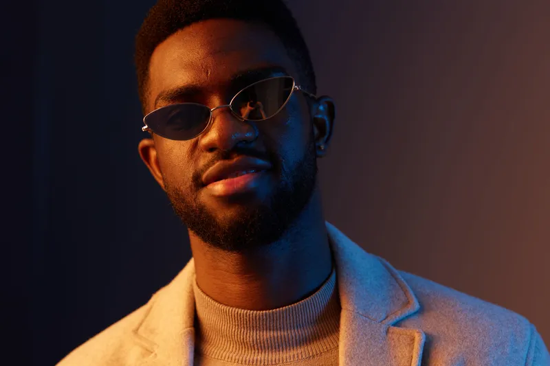 Stylish young Black man wearing sunglasses, exuding confidence, set against a colorful, gradient background that highlights modern fashion and urban aesthetics