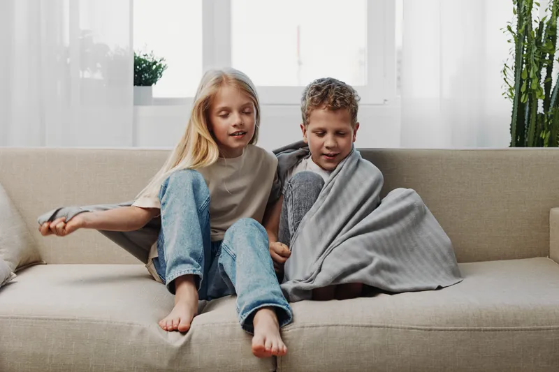Two children sitting on a sofa, one wrapped in a cozy blanket, enjoying a relaxed moment in a bright living room.