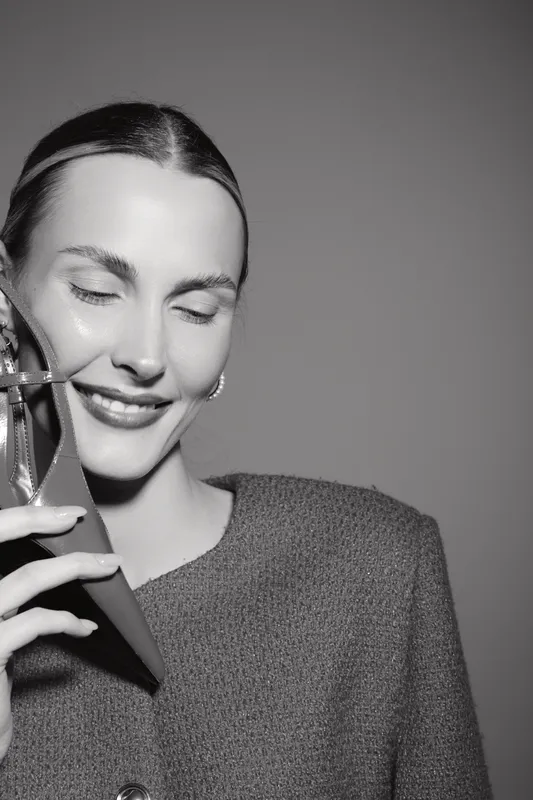 Smiling white woman with light skin and brunette hair holds high heel shoe near face. Studio portrait in black and white with joyful expression. Concept of fashion and happiness.