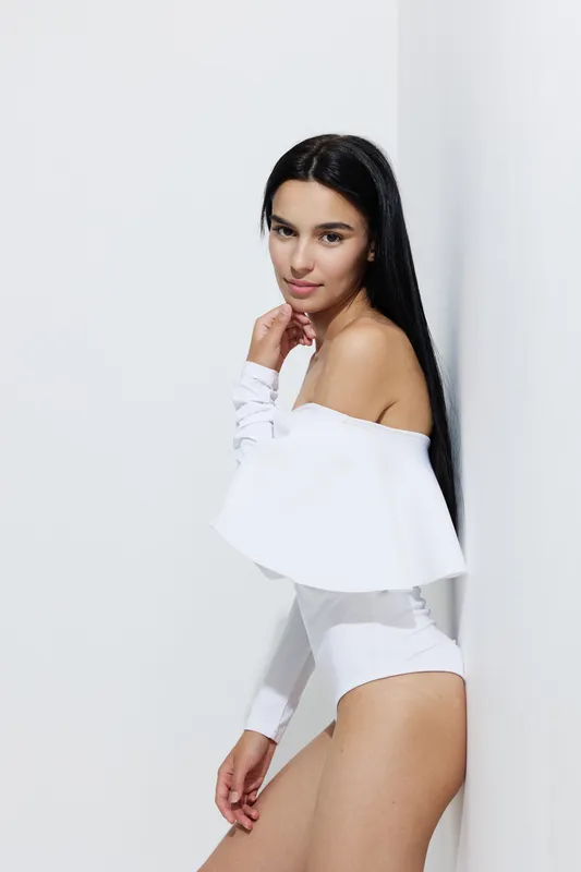 Sport fitness woman posing indoors with long dark hair and white activewear, looking confident and focused in a minimalistic bright studio environment.