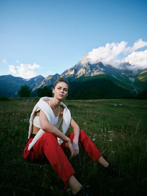 Woman sitting on a meadow grass in redpants and a light knit top against a vast mountain landscape, clear sky and open fields, showcasing outdoor fashion, calm mood, and nature inspired scenery