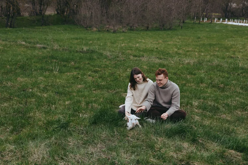 Young couple sitting on green grass in a park playing with a small white baby goat during a casual outdoor lifestyle moment in cool weather.