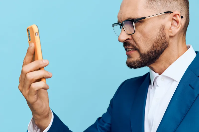 A professional man in a suit checks his phone with focused emotion against a solid blue background, conveying confidence, concentration, and modern business readiness.