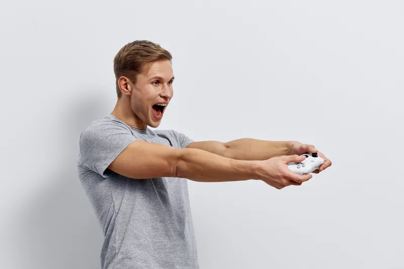 Enthusiastic young man with short brown hair playing video game, holding game controller with both hands, excited facial expression, standing indoors against plain white background, casual grey t-shir