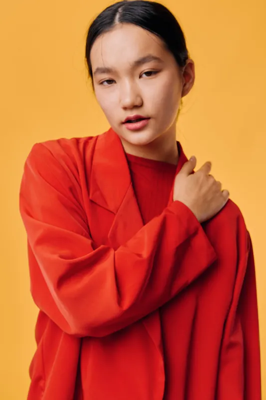 Young woman with black hair wears red oversized coat standing against yellow background with hand on shoulder looking confidently at camera in bright studio portrait.
