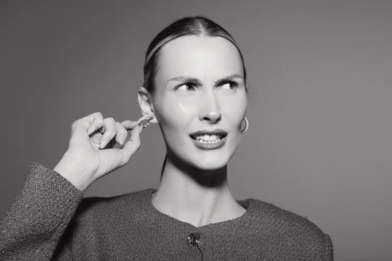 Portrait of a young Caucasian woman with light skin pulling her ear and grimacing, wearing earrings and a textured top on a grey background.