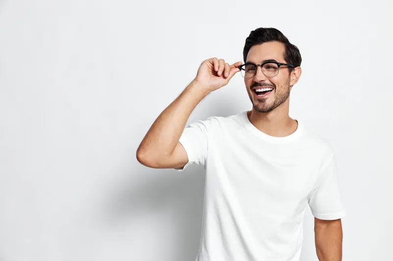 Smiling man wearing glasses and a white t-shirt looks at the camera with a cheerful expression while holding his glasses. Isolated on plain light background.
