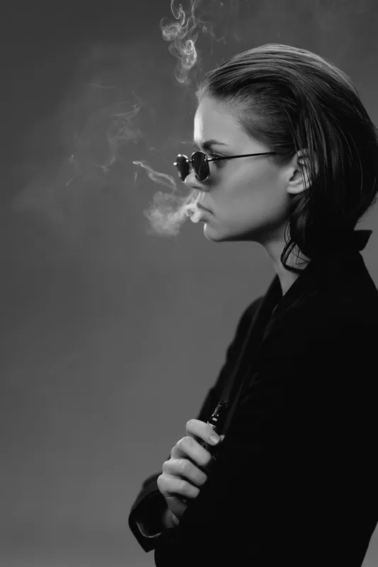 Black and white portrait of a stylish woman smoking in sunglasses with a moody atmosphere and dark background