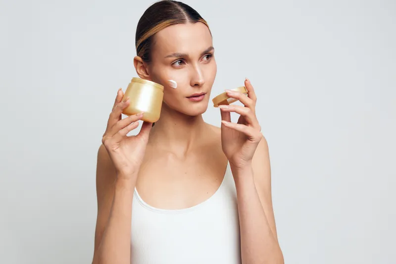 Young woman applying cream on face holding open jar with skincare product focused on beauty routine natural clean skin white tank top studio background.