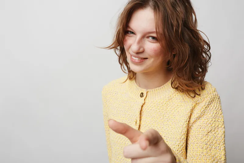 Smiling young woman with curly hair wearing yellow sweater points finger at camera. Studio portrait on grey background. Concept of confidence, friendliness, and positive attitude.