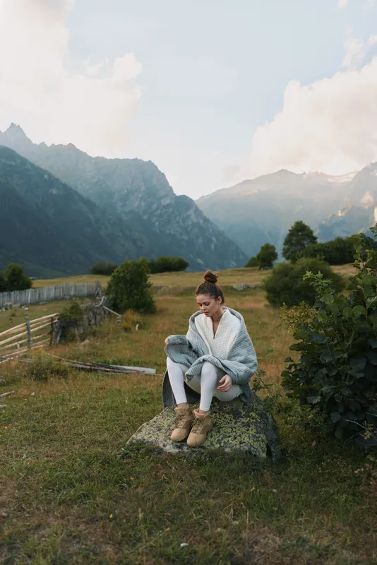 Young woman sits on a rock in a mountain meadow, wrapped in a light cardigan, looking thoughtful as distant valley and towering peaks rise behind.