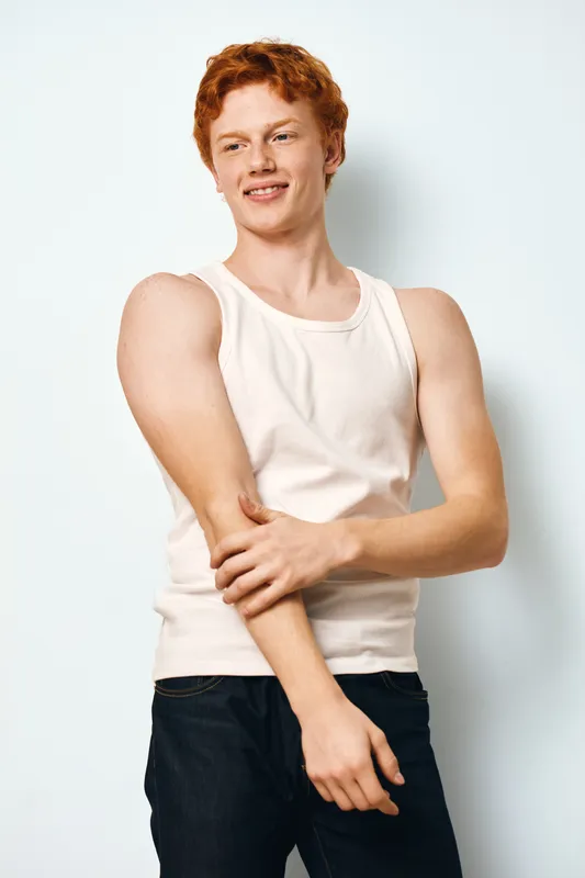 Young man with red hair wearing white tank top and black jeans standing against white background with relaxed posture and subtle smile.
