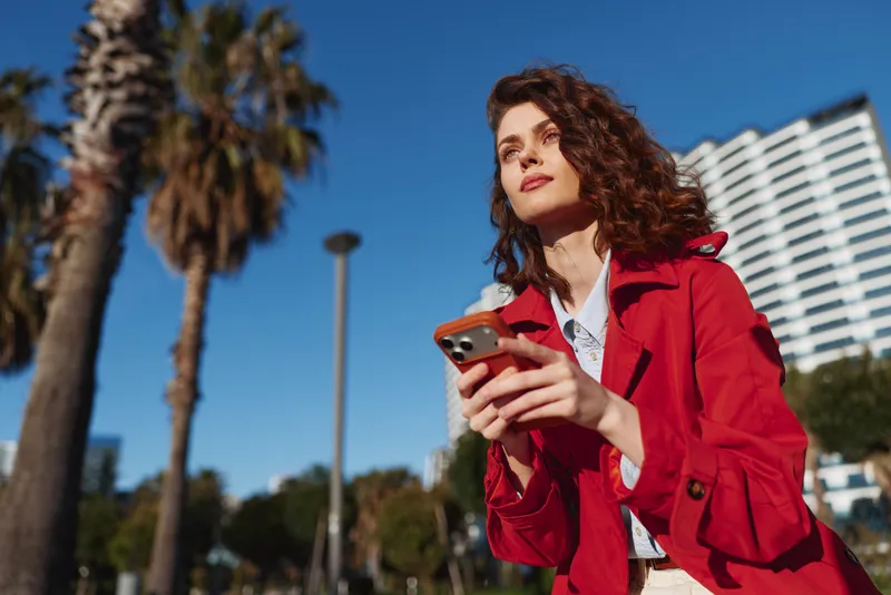 Young woman in a red trench coat stands in an urban outdoor setting, holding a smartphone with a confident expression, curly brown hair, palm trees and blue sky behind.