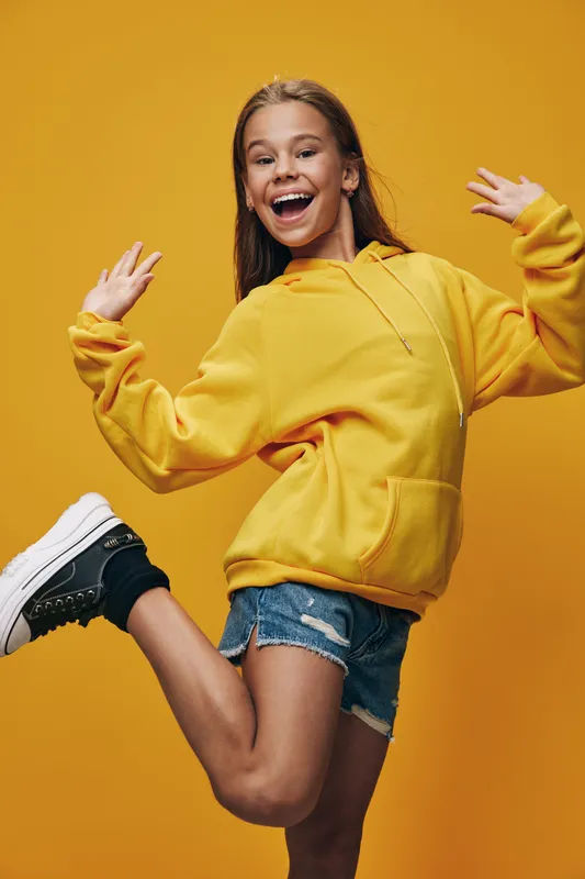 Happy teenage girl posing on one leg wearing yellow hoodie and denim shorts with casual shoes against yellow background for youth fashion and lifestyle.