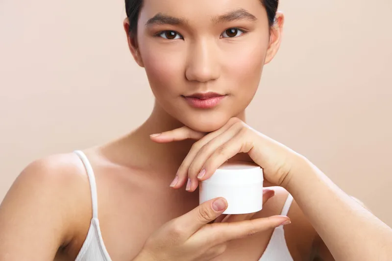 asian woman holds a small cream jar near her face in a soft studio setting, highlighting skincare focus. her calm expression and gentle lighting convey a serene, healthy beauty moment for consumers