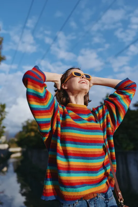Woman in colorful striped sweater and sunglasses smiling outdoors, relaxed portrait with hands behind head, casual fashion and positive mood under bright blue sky.