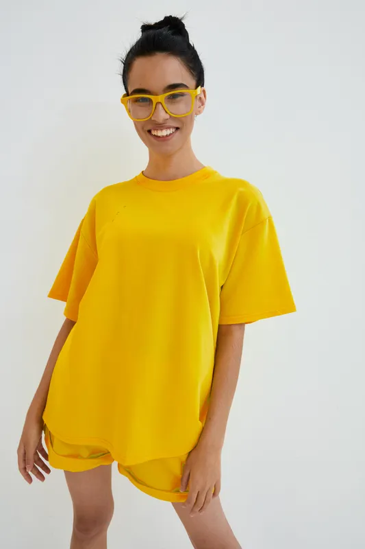 smiling woman wearing bright yellow casual outfit and matching eyeglasses standing against white background with cheerful expression and natural hairstyle