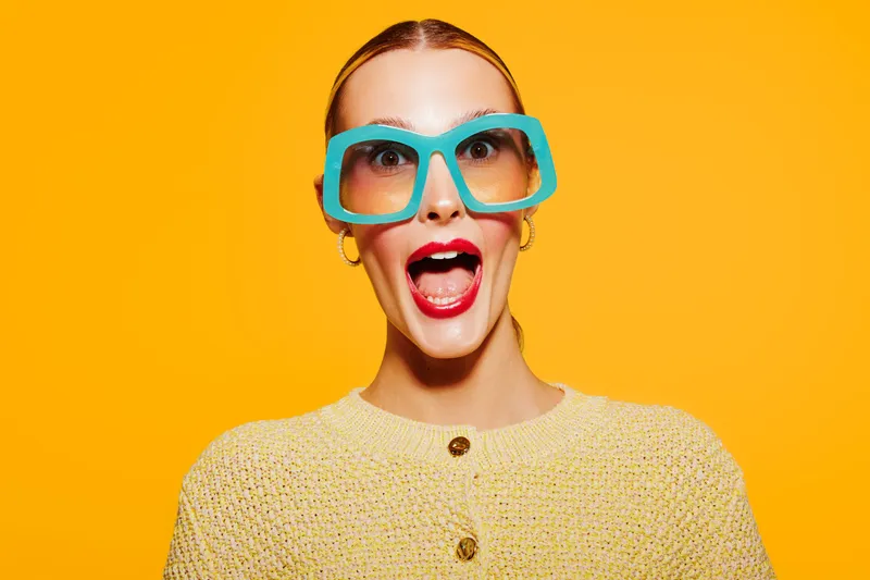 Surprised young woman with bright red lipstick and large blue sunglasses on yellow background. She wears a textured beige sweater and expresses excitement or shock with open mouth.