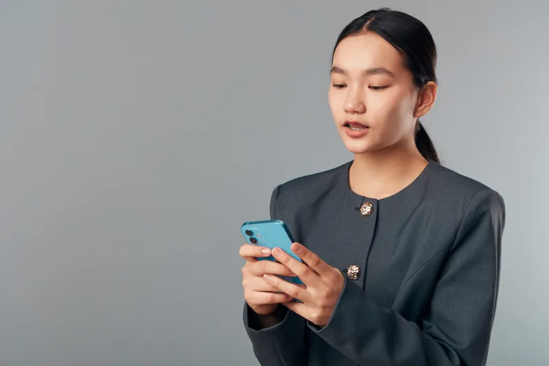Young woman holding mobile phone looking at screen thoughtfully in studio with plain background, technology and communication concept for online connection or social media.