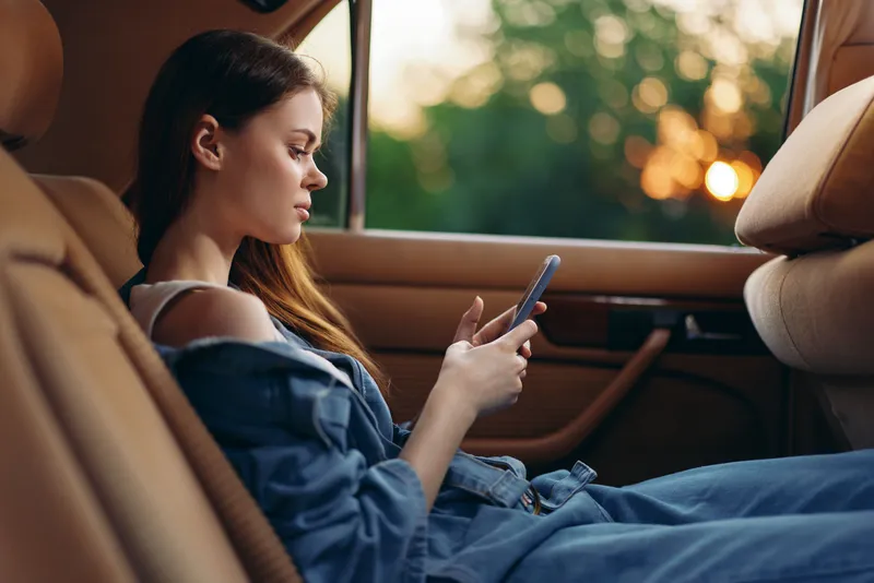 Young woman in denim outfit using smartphone while sitting in a car during sunset, capturing relaxed mood and urban lifestyle.