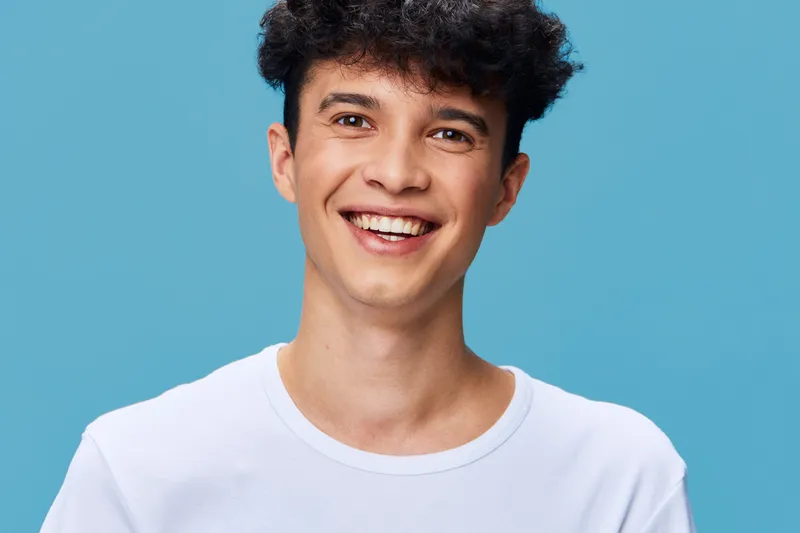 smiling man with curly hair wearing a white t-shirt stands against a vibrant blue background, radiating happiness and youthfulness in a casual studio portrait.