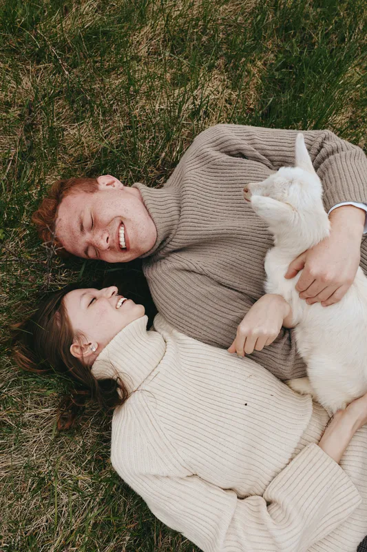 A relaxed couple rests on grass, smiling and cuddling a white goat kid. Outdoors lifestyle moment capturing affection, calm, and bonding with a charming farm animal.