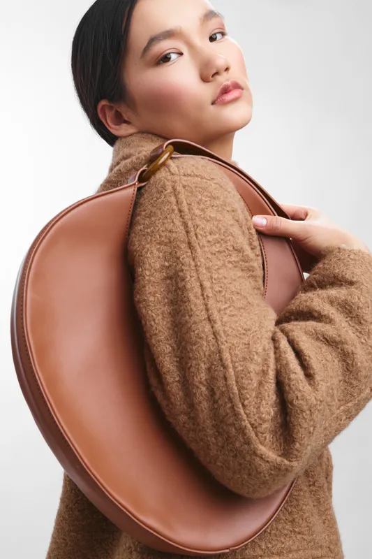 asian woman wearing a camel coat with a round brown bag over her shoulder, exudes modern elegance and confidence, lighting highlighting her features against a clean backdrop.