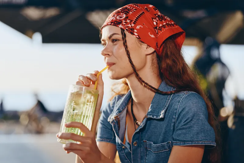 Young woman with red bandana drinking a refreshing cold beverage outdoors in warm sunlight, casual style and relaxed mood in summer setting.