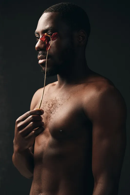 Black man with bare chest holding dried flower near face in creative conceptual visual style, artistic portrait with dark background and thoughtful expression in studio lighting.