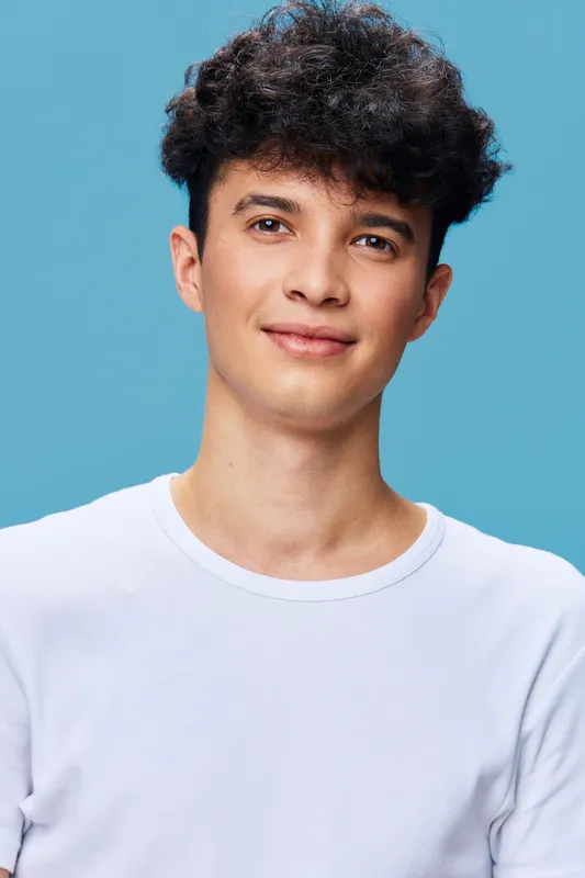 young man with curly hair wearing white shirt smiles gently against pastel blue background, studio portrait, casual style, natural lighting